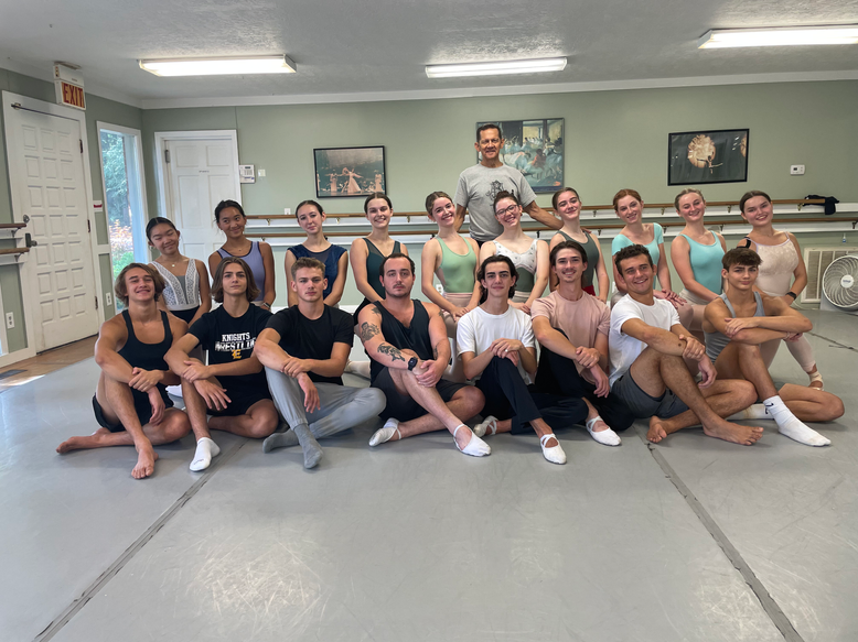 A group of ballet dancers sitting on a studio floor, posing for a group photo with their instructor standing behind them.