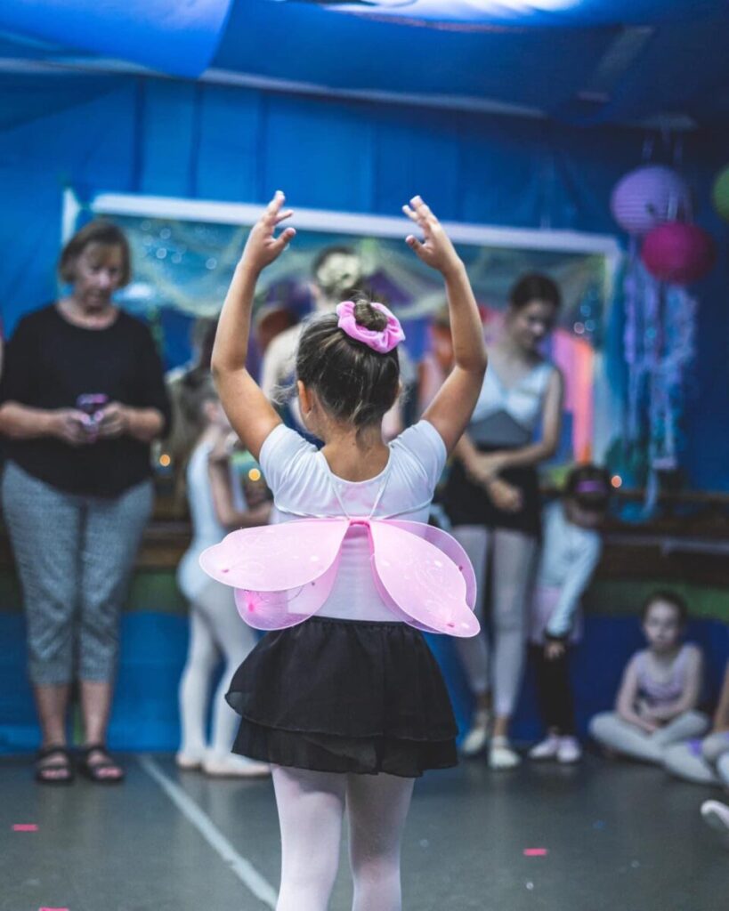 A young girl in ballet attire with butterfly wings stands posed in a dance studio, while others watch in the background.